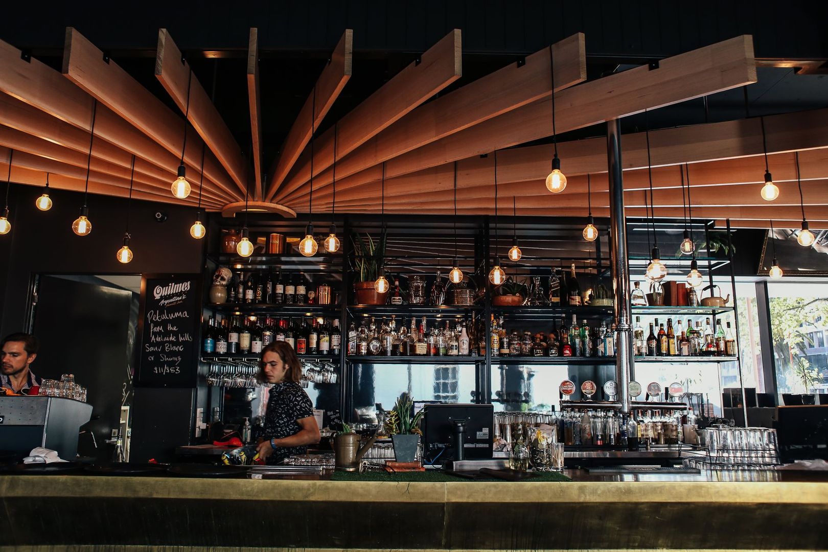 A modern cocktail bar interior with wooden ceiling panels and hanging bulbs